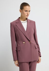 Dubbelzijdige blazer van mauve stof met geknipte revers, gouden knoopaccenten en zijzakken. Draagt over een witte top met versieringen.