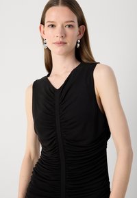 Černé bezrukávové šaty s řasenou strukturou, véčkovým výstřihem a přiléhavým střihem. Obsahují tónované švy a hladkou látku. Minimalistický design.