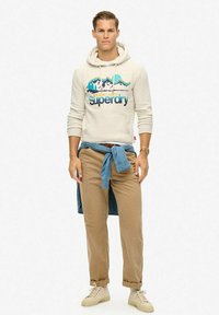 Beżowa bluza z kapturem z graficznym motywem gór, niebieskimi akcentami i napisem "Superdry". Jasnobeżowe spodnie i jasnoniebieska koszula przewiązana w talii. Beżowe sneakersy.