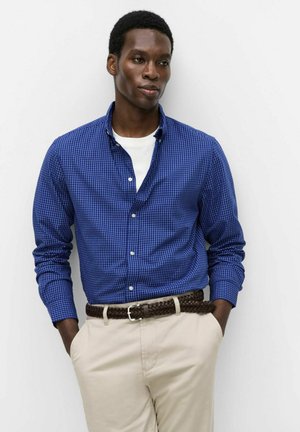 Man draagt een blauw geruit overhemd over een wit T-shirt, beige broek en een bruine gevlochten riem met handen in de zakken.