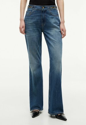 Vaqueros azules de pierna ancha con bolsillos delanteros y pequeñas trabillas decorativas, combinados con zapatos negros puntiagudos y una camiseta negra.