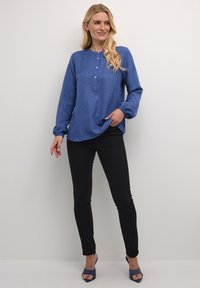 Blauwe blouse met lange mouwen van gestructureerde stof en knoopdetails, gecombineerd met zwarte skinny jeans en blauwe open die schoenen. Moderne, casual look.