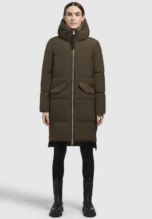 Žena stojaca v dlhom olivovo zelenej prešívanej zimnej bunde s vysokým golierom, čiernych legínach a čiernych členkových topánkach na jednoduchom pozadí.