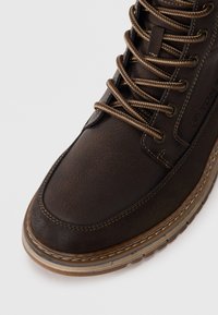 Brun læder høj-top sko med struktureret overflade, kontrasterende brune snørebånd og beige gummisål. Har syninger og brandede detaljer.