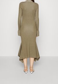 Femme portant une robe longue à manches longues en tricot côtelé taupe avec un ourlet asymétrique évasé et des sandales à talons bas blanches à brides, de dos.