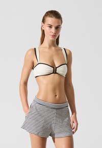 Top de bikini en color crema con ribete negro y detalle de nudo frontal, combinado con shorts de cintura alta con un patrón geométrico en blanco y negro.