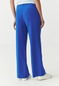 Niebieskie spodnie o szerokich nogawkach wykonane z gładkiego materiału, z talią na wysokości połowy, tylnymi kieszeniami oraz plisami wzdłuż nóg.