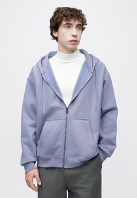 Casaco com capuz de cor lilás claro com fecho de correr, capuz grande, bolsos frontais e corte descontraído; combinado com uma gola alta branca e calças cinza.