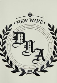 Svart broderad logotyp med "NEW WAVE DNA 1998" inuti en cirkulär design omgiven av lagerblad på en ljus färgad tygbakgrund.