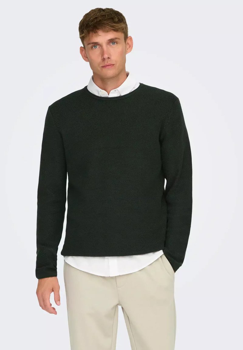 Maglione a maglia verde scuro con scollatura a girocollo, tessuto testurizzato, maniche lunghe e fit rilassato, indossato sopra una camicia bianca.