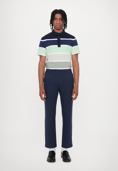 Polo skjorte med marineblå, grønn og hvite striper; kombinert med marineblå bukser; svarte loafers; ensfarget hvit bakgrunn.