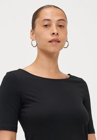 T-shirt nera aderente con colletto rotondo e maniche corte, realizzata in un tessuto morbido. Modello che indossa orecchini a cerchio in argento.