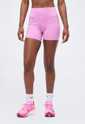 Personne athlétique portant un short ajusté rose clair, des chaussettes blanches Nike et des chaussures de course rose vif, debout sur un fond blanc.