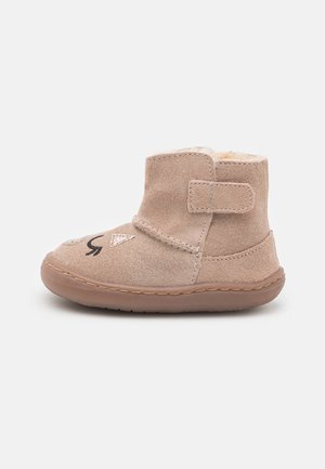 Botas de tobillo de ante color beige con un suave forro de piel, que cuentan con una correa de velcro y detalles bordados de una cara en la parte frontal. Suela de goma plana.