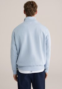 Šviesiai mėlynas džemperis su aukštu apykakliu, siaurėjančiomis rankogaliais ir apačia. Minkšta medžiaga su minimalistišku dizainu, matomomis siūlėmis ir sluoksniuotu išvaizda.