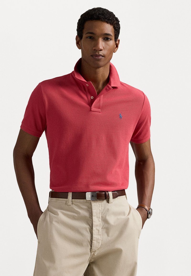 Κόκκινο κοντομάνικο πόλο μπλουζάκι φτιαγμένο από βαμβάκι, με διπλό κουμπί και μικρό κεντημένο λογότυπο. Συνδυάζεται με χακί παντελόνι και ζώνη.