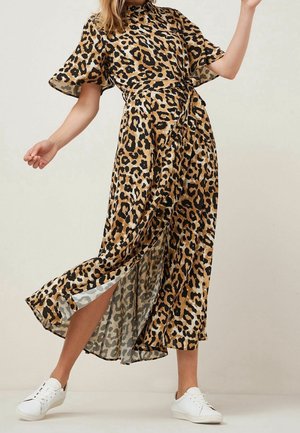 Frau trägt ein fließendes Midikleid mit Leopardenmuster und Flatterärmeln sowie weiße Sneaker, posiert mit einem Bein, das durch einen seitlichen Schlitz sichtbar ist.