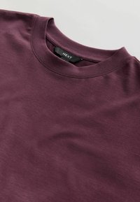 Bordo värvi ümarakaelusega T-särk, valmistatud pehmest kangast, sileda tekstuuriga; krae lähedal on silt, millel on kiri "NEXT".