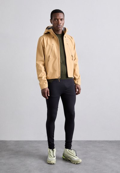 Veste imperméable marron clair avec capuche, deux poches avant zippées, associée à un pantalon noir ajusté et des chaussures vert clair.