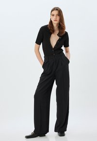 Bodysuit negro de canalé con escote en V profundo y mangas cortas, combinado con pantalones negros de cintura alta con pleats marcados y un corte entallado.