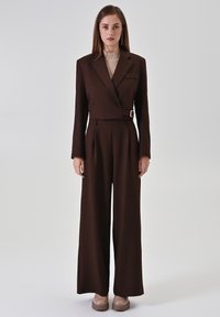 Bruin op maat gemaakte pak met een double-breasted jas en broek met wijde pijpen. Heeft een gespdetail aan de taille en een beige geribbelde trui eronder.