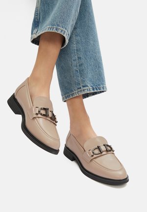 Beżowe skórzane mokasyny z ozdobną klamrą, niską czarną podeszwą i przeszyciami, sparowane z podwiniętymi niebieskimi dżinsami.