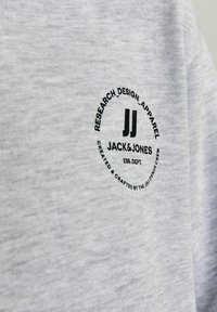 Tricou din bumbac gri cu un logo rotund negru ce conține textul "JACK & JONES" și detalii. Materialul are un aspect moale și texturat.