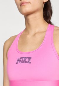 Roze sportbeha met een ronde halslijn en brede banden, met een opvallend zwart "NIKE"-logo gecentreerd op de borst. Gladde textuur.