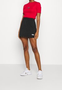 Červená košile s krátkým rukávem a logem, kombinována s černou sukní se zipem vpředu. Bílé tenisky dokreslují celý outfit a zdůrazňují ležérní styl.