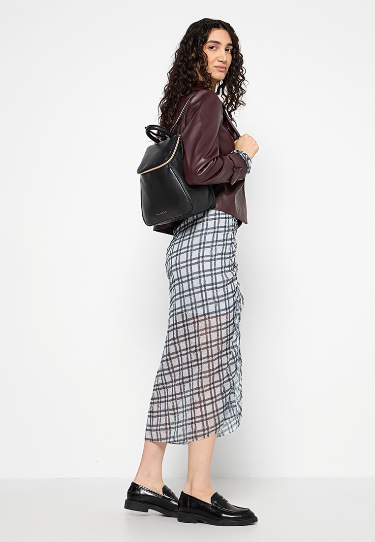 Un sac à dos en cuir noir avec une fermeture à glissière dorée, porté sur une veste bordeaux et une jupe midi en plaid et transparente. Des mocassins noirs complètent la tenue.