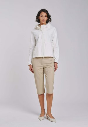 Vrouw met kort donker haar die een witte hoodie met ritssluiting draagt, beige knielange broek en witte pumps met punten, staand tegen een effen achtergrond.