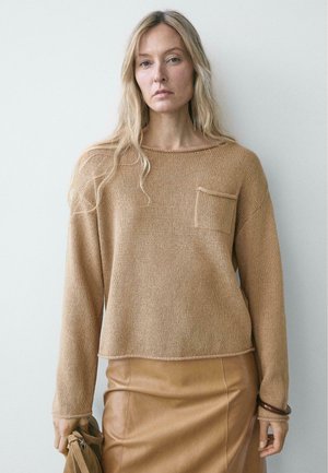 Femme blonde portant un pull en maille camel ample avec une poche poitrine et une jupe en cuir beige, tenant un sac assorti devant un fond uni.