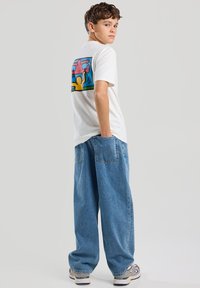 Valkoinen puuvillapaita, jossa on värikäs grafiikka selässä esittäen abstrakteja hahmoja. Yhdistetty löysään, sinisiin farkkuihin.