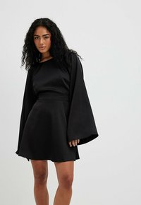 Robe noire avec de longues manches cloche, texture satin lisse, taille cintrée et jupe évasée. Encolure ronde simple, sans motifs ni accents visibles.