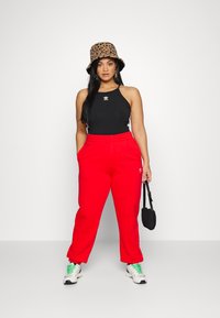 Pantaloni della tuta rossi, top halter nero con accenti bianchi, cappello a secchiello con stampa leopardata e sneakers bianche con lacci verdi. Borsa nera in mano.