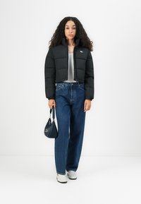 Black cropped puffer jacket with a zipper, gray top, wide-leg blue denim jeans, and white sneakers. Small black handbag with strap.