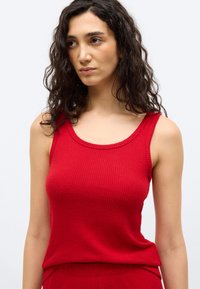 Γυναίκα με μακριά σγουρά σκοτεινά μαλλιά που φοράει μια αμάνικη κόκκινη ριμπ μπλού ζα, στέκεται μπροστά σε απλό ανοιχτό φόντο και κοιτάζει ελαφρώς προς τα δεξιά.