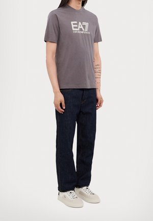 Personne portant un t-shirt gris avec le logo Emporio Armani EA7, un jean bleu foncé et des baskets blanches, debout devant un fond neutre.