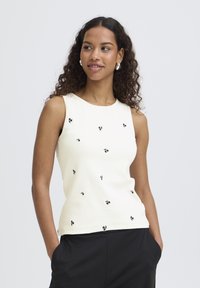 Giovane donna con capelli ricci che indossa una canottiera bianca con piccoli motivi floreali neri e pantaloni neri, con le mani in tasca, che guarda di lato.