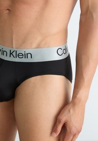 Schwarze Herrenunterhosen mit einem silbernen elastischen Bund, der das Logo "Calvin Klein" trägt. Glattes Material mit einer körpernahen Passform und minimaler Gestaltung.