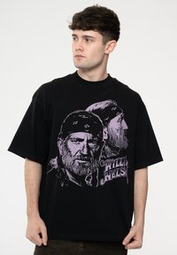 Black cotton t-shirt featuring a graphic of two faces and the text "WILLIE NELSON" in purple. Short sleeves and relaxed fit.