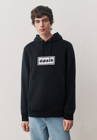 Zwarte hoodie met trekkoorden, voorzien van een prominent wit rechthoekig logo met de tekst "oasis" in het midden van de borst.