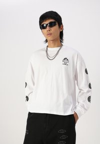 White long-sleeve shirt in cotton, featuring black graphic prints on sleeves and front, paired with black pants and silver layered chains.