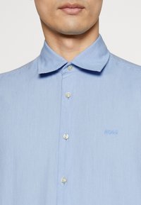 Primer plano de una persona que lleva una camisa de botones azul claro con cuello y un pequeño logotipo "BOSS" bordado en el pecho.
