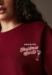 Bordo krāsa sporta krekls ar baltiem un rozā izšūtiem uzrakstiem: "Ievadīsim Ziemassvētku dzērienus" un martini glāzes grafiku. Mīksta, kokvilnai līdzīga tekstūra.