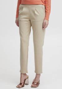 Pantalons beige ajustés avec une texture lisse, avec des plis avant et des fentes latérales. Assortis à des mules transparentes à talons.