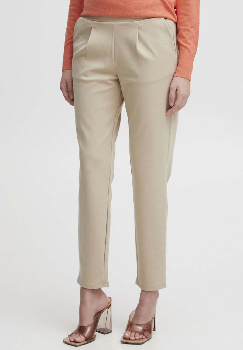 Pantalons beige ajustés avec une texture lisse, avec des plis avant et des fentes latérales. Assortis à des mules transparentes à talons.