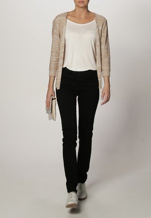 Cardigan en tricot beige sur un t-shirt blanc, associé à un jean skinny noir et des baskets blanches. Tenant une pochette de couleur claire.