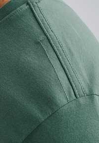 Tessuto di cotone verde che mostra un primo piano dei dettagli di cucitura e della texture, con cuciture che scorrono lungo l'area delle spalle e una finitura liscia.