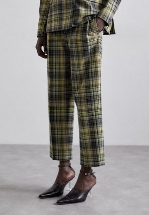 Person trägt grüne und schwarze karierte, kurze Hosen und passendes Oberteil, mit schwarzen spitzen Schuhen mit transparenten Blockabsätzen.